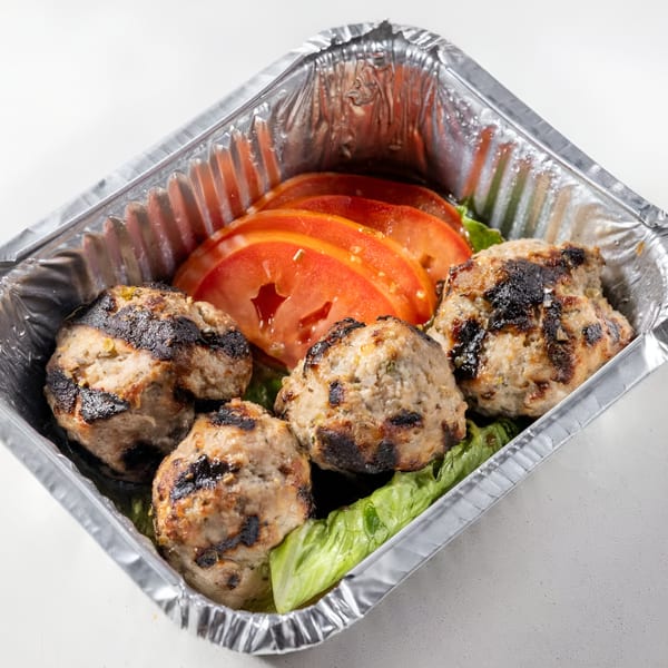 Grilled Greek meatballs with charred marks, sliced tomatoes, and lettuce in a foil takeout container.